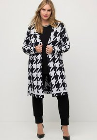 Zwart-wit houndstooth patroon lange cardigan met knoopsluiting, gecombineerd met een zwart top en slanke broeken, afgewerkt met zwarte hakken.