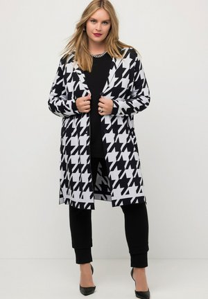 Dlouhý cardigan s černobílým vzorem houndstooth a zapínáním na knoflíky, kombinovaný s černým topem a úzkými kalhotami, doplněný černými lodičkami.