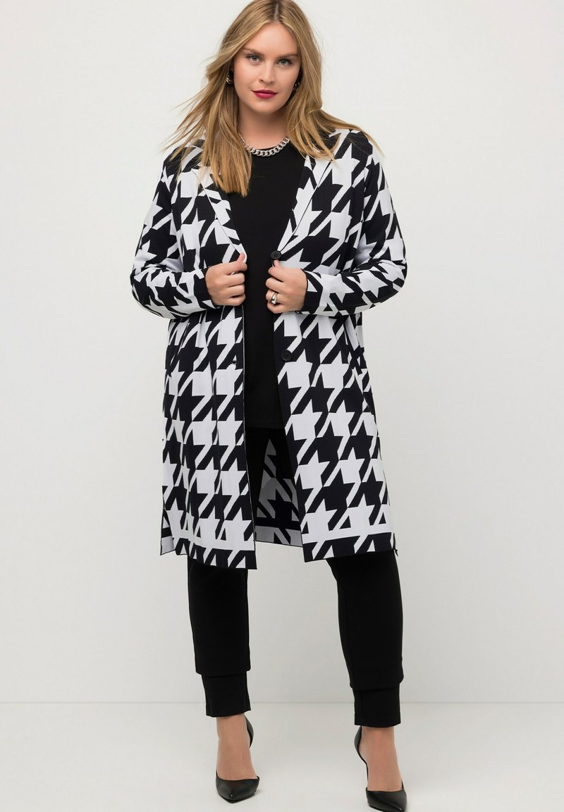Zwart-wit houndstooth patroon lange cardigan met knoopsluiting, gecombineerd met een zwart top en slanke broeken, afgewerkt met zwarte hakken.