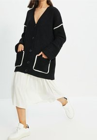 Svart stickad cardigan med djup V-ringning, stora knappar och vita detaljer längs fickorna och ärmarna, bärs över en vit kjol.