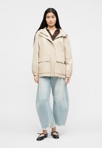 Femme portant une veste à capuche beige, un chemisier marron foncé, un jean large bleu clair aux ourlets effilochés et des ballerines noires à brides.