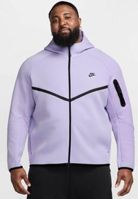 Felpa con zip Nike viola con accenti neri, dotata di colletto alto, tasca con zip sulla manica e cuciture della spalla a contrasto. Materiale morbido.