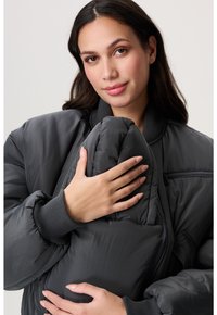 Frau mit langen, dunklen Haaren, die eine dunkle Daunenjacke trägt und sanft einen Teil der Jacke nah an ihre Brust hält, neutraler Hintergrund.