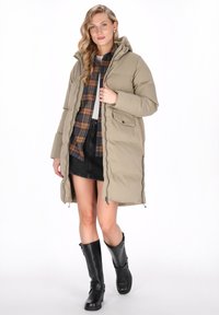 Vrouw in een beige gewatteerde jas met capuchon over een geruite shirt, zwarte rok en zwarte kniehoge laarzen, staand tegen een witte achtergrond.