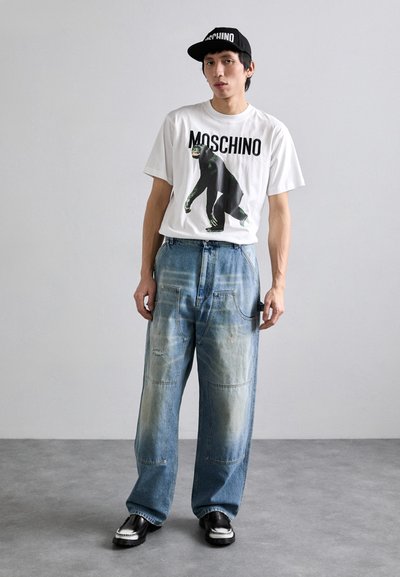 Fehér póló fekete grafikai nyomtatással egy gorilláról, kék bőszárú farmer, amelyen kopott részletek vannak, valamint fekete sapka "MOSCHINO" logóval.