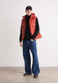 Rød polstret vest med hette over en svart genser med høy hals, kombinert med blå vide bukser og grå sko. Enkelt design, uten synlige mønstre.
