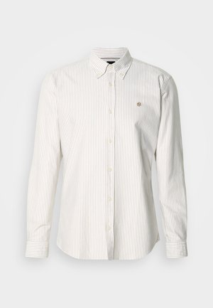 Világos bézs és fehér csíkos hosszú ujjú ing. Gallérja gombos, elöl gombokkal záródik, és a mellrészen egy kis logó található.