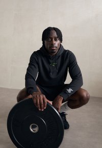 Melnā sporta jaciņa ar zaļu "HYROK" tekstu, melni šorti, turot melnu svaru plati pelēkā grīdā; teksturēts fons.