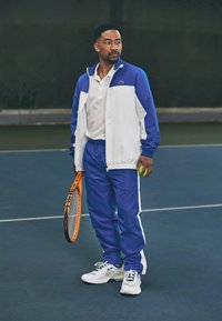 Zils un balts sporta tērps ar r�āvējslēdzēja jaku, saskaņotiem biksēm un baltām sporta kedām, tur rokās oranžu tenisa raketi un tenisa bumbu.