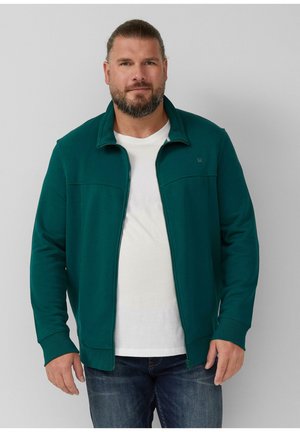 Homme barbu avec les cheveux attachés, portant une veste zippée teal sur un t-shirt blanc et un jean bleu, se tenant devant un fond uni.
