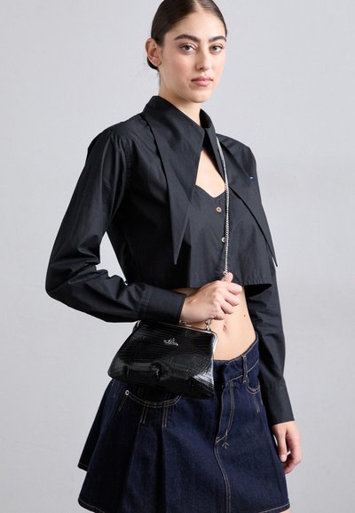 Tânără îmbrăcată într-un tricou scurt negru și o fustă din denim închis, ținând o geantă mică neagră cu o curea de lanț argintie.