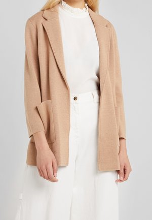 Frau trägt einen beigen Blazer mit offenem Vorderteil über einer weißen Bluse mit Rüschenkragen und hoch taillierter weißer Hose mit Knopfdetail.