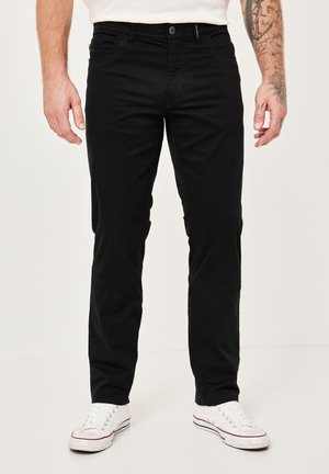 Schwarze, figurbetonte Hosen mit vorderen Taschen und einem Reißverschluss, aus einem Baumwollgemisch. Getragen mit weißen Sneakers in niedriger Form.