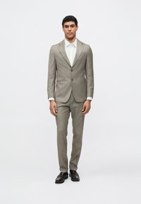 Jasnobrązowy, dopasowany garnitur w kratę, zestawiony z białą koszulą. Spodnie o standardowym kroju. Całość dopełniają brązowe skórzane buty.