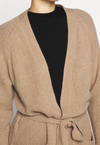 Beżowy kardigan wykonany z miękkiego, teksturowanego materiału z głębokim dekoltem w serek, fasonem owijanym i przewiązanym w pasie, z dwoma przednimi kieszeniami.
