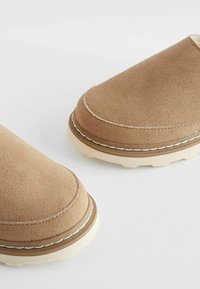 Brown suede slip-on shoes with a rounded toe, light-colored rubber sole, and contrasting stitching detail around the upper edges.