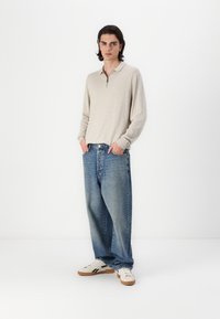 Beige langærmet polo skjorte med lynlås, parret med løse blå denim jeans og hvide sneakers med sorte detaljer. Ensfarvet baggrund.