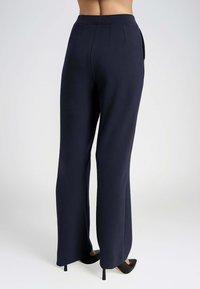 Pantaloni a gamba larga blu navy con una texture liscia, caratterizzati da pieghe frontali e tasche laterali, abbinati a tacchi a punta neri.