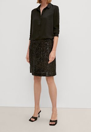 Femme portant un chemisier noir à boutons, une jupe noire à sequins longueur genou et des mules à talons hauts ouvertes à l'avant, debout devant un fond uni.