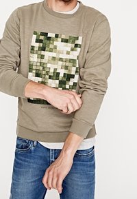 Felpa verde oliva con una grafica pixelata verde e bianca sul davanti, dotata di polsini e orlo a coste, indossata con jeans blu.