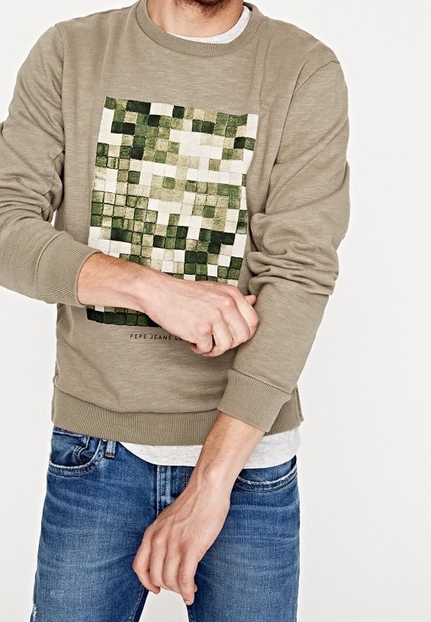 Felpa verde oliva con una grafica pixelata verde e bianca sul davanti, dotata di polsini e orlo a coste, indossata con jeans blu.