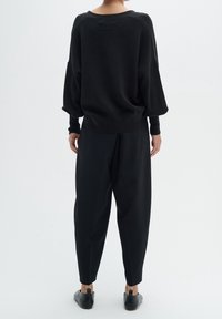 Černý oversized svetr se širokým výstřihem, kombinovaný s volnými černými kalhotami a černými botami na nízkém podpatku, viděný zezadu.