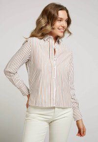 Chemise blanche avec de fines rayures verticales marron, design à boutons, manches longues avec poignets à boutons, et un ourlet légèrement arrondi.