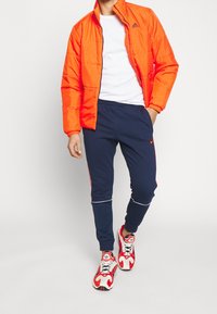 Světle oranžová vycpaná bunda s vysokým límcem, bílý tričko, námořnické tepláky s červenými postranními pruhy a červené a bílé sportovní boty.