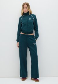 Chaqueta de color teal con cremallera y ribete blanco, combinada con pantalones de chándal a juego con cintura elástica. Ambos confeccionados en un suave tejido texturizado.