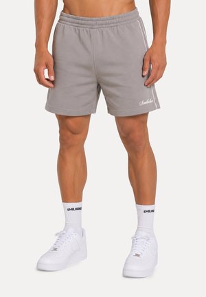 Man draagt grijze sportshorts, witte sokken met de tekst "SMILODOX" en witte sneakers, staand tegen een effen achtergrond.