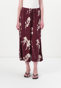 Burgunderfargede culotte bukser med kremfarget blomsterdesign, elastisk midjebånd med snøring og vide ben. Kombinert med svarte flip-flops.