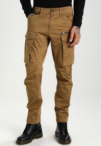 Cargo-housut khaki-värissä, valmistettu kestävästä kankaasta. Ominaisuuksiin kuuluu useita taskuja, sopiva vyötärö ja kapenevat lahkeet. Yhdistetty mustiin saappaisiin.