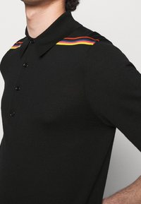 Czarny polo z guzikami, z kolorowymi poziomymi paskami w czerwonym, żółtym i niebieskim na ramionach. Miękka tekstura, dopasowany krój.