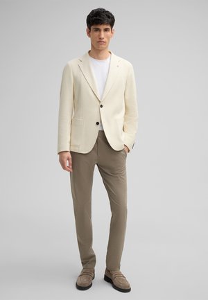 Hellbeige strukturierter Blazer mit zwei Vorder Taschen, schwarzen Knöpfen, kombiniert mit hellbraunen Hosen und grauen Slippern. Darunter ein einfaches T-Shirt mit Rundhalsausschnitt.
