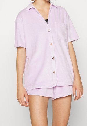 Fliederfarbenes Kurzarmhemd aus weichem Stoff, mit Knopfleiste, Kragen und einer einzelnen Brusttasche. Kombiniert mit passenden Shorts.