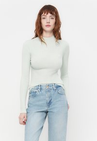Lys grønn ribbet langermet topp med høy hals, sammen med lys blå høyhalsede jeans. Har en liten logo på brystet.