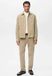 Beige corduroy jacket with two chest pockets, paired with matching beige corduroy pants and dark shoes, featuring a white turtleneck underneath.