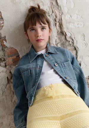 Giovane con capelli castani, indossa una giacca di jeans e una maglietta bianca, in piedi davanti a una parete texturizzata e rovinata dal tempo.