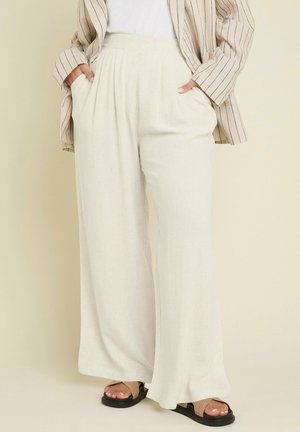 Person iført løstsiddende, cremefarvede bukser med vide ben og sidelommer, kombineret med en beige blazer med striber og sorte sandaler på en lys baggrund.