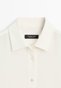 Bílá halenka s límečkem, zapínáním na knoflíky vpředu, jemnou texturou a štítkem s nápisem "Massimo Dutti" u výstřihu.
