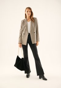 Kvinde stående iført en ternet blazer, hvid top, sorte brede jeans, sorte støvler, holder en stor sort håndtaske med en guldkæde som strop.