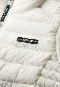 Weißer Puffer-Jacke mit gestepptem Muster, ausgestattet mit einem schwarzen Superdry-Logo-Patch und einem hellblauen Reißverschlussakzent. Glattes, elegantes Design.