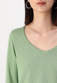 Maglietta verde a maniche lunghe con scollo a V, realizzata in tessuto morbido e testurizzato. Modello abbinato a grandi orecchini circolari.