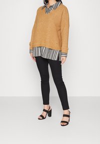 Sinapinkeltainen neulepaita V-pääntiellä mustavalkoraidallisen paidan päällä, yhdistetty mustiin leggingseihin ja mustiin remmiblokkitakakorkoisiin sandaaleihin.