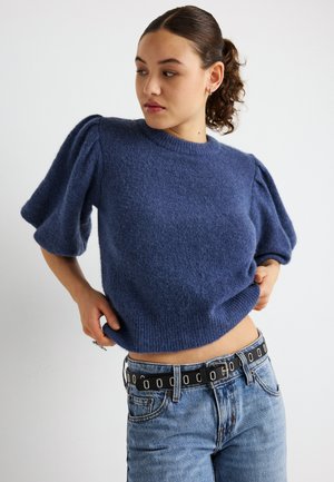 Blauer Strickpullover mit gepufften kurzen Ärmeln, rundem Ausschnitt und strukturierter Oberfläche. Kombiniert mit hoch taillierten Jeans und einem schwarzen Gürtel.