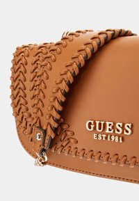 Ruda odinė rankinė su pintinėmis detalėmis, auksinės spalvos apdaila ir iškilniu logotipu "GUESS EST 1981" priekyje.