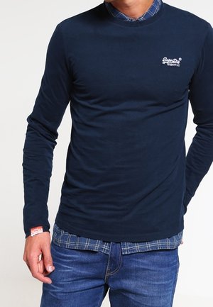 Mężczyzna w granatowej koszuli z długim rękawem ubranej na niebieską koszulę w kratę na guziki oraz niebieskie dżinsy, stoi z jedną ręką schowaną za plecami.