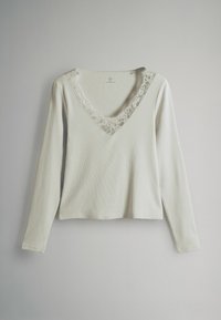 Dlhý rukáv s výstrihom do V v svetlosivej rebrovanej látke; čipkový lem pozdĺž výstrihu pridáva textúru a detail. Žiadne viditeľné zapínanie.