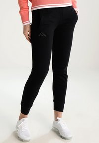 Person som bär svarta slim-fit joggers med sidfickor och vita sneakers, stående med händerna i fickorna.