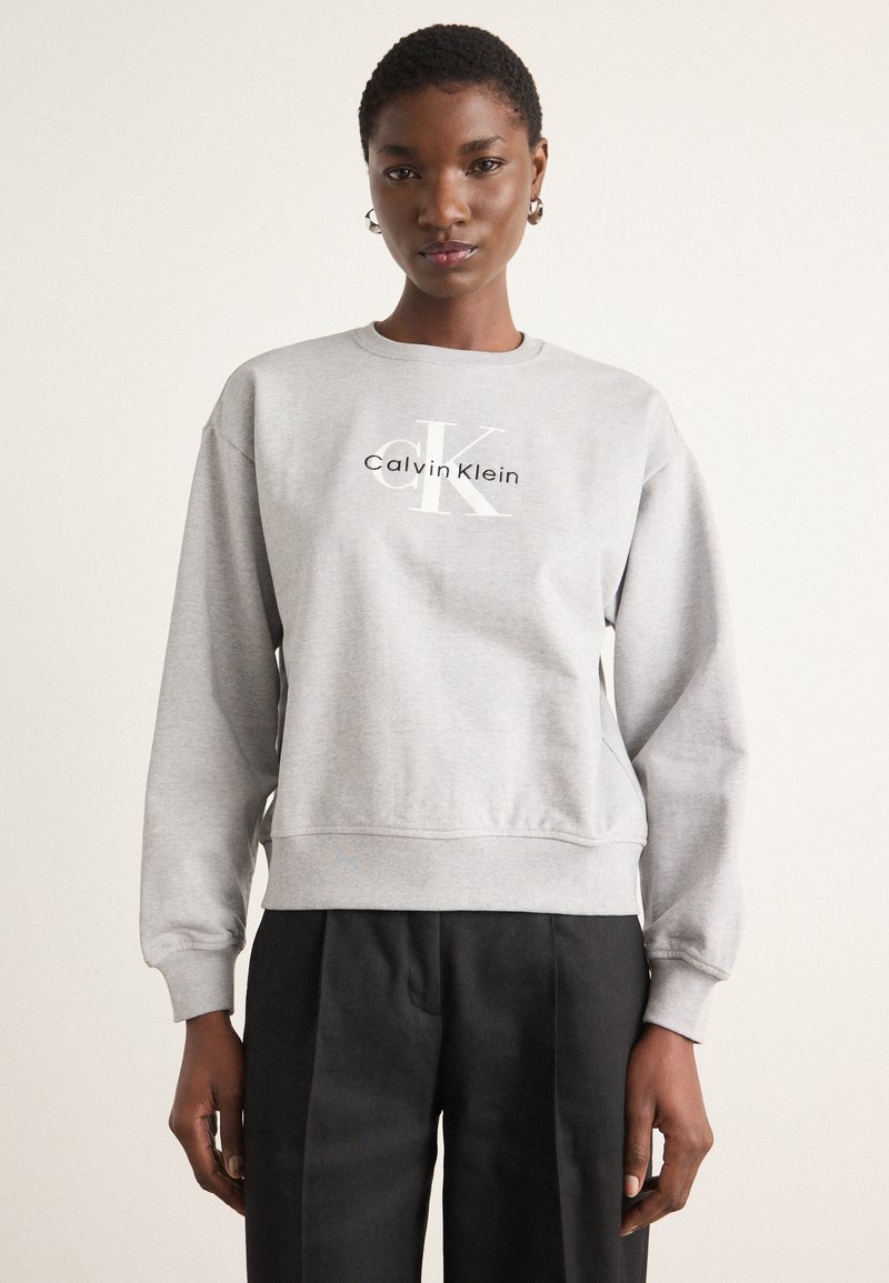 Gray sweatshirt made of soft fabric, featuring a round neckline and ribbed cuffs, with a logo in white across the chest. Paired with black trousers.
